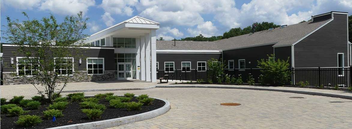Modern building with a slanted roof and large windows set against a blue sky. The facade is a mix of dark siding and stonework. A roundabout with paved pathways and landscaped with low shrubs is in front.
