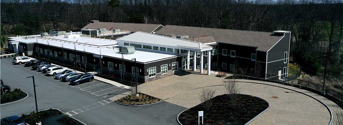 Aerial view of a large, single-story building with a mix of white and dark exterior. The building has a flat roof and a prominent entrance with columns. A parking lot is adjacent, with several parked cars and visible landscaping. Trees surround the area.