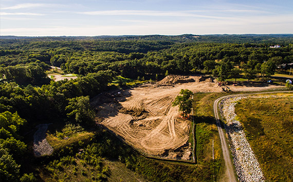 Aerial view of a construction site surrounded by dense green forest. The cleared land has visible dirt tracks and mounds of earth. A road curves along the right side, and hills are visible in the distant background under a clear sky.