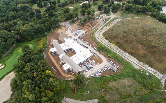 Aerial view of a large construction site surrounded by trees. The site includes a rectangular building under construction, with dirt roads and parked vehicles around it. Adjacent is a grassy area with exposed soil and a golf course nearby.