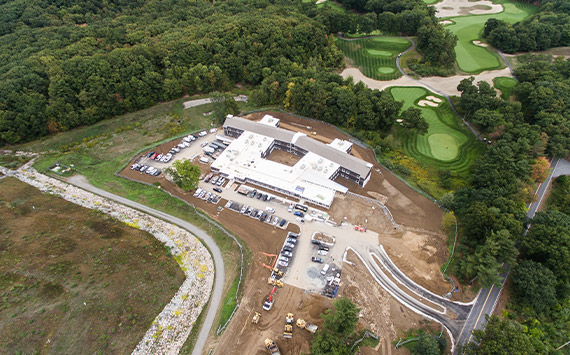 Aerial view of a construction site surrounded by a forest and golf course. A large building is partially surrounded by parked cars and construction machinery. Nearby, neatly maintained golf greens and sand traps are visible amidst the trees.