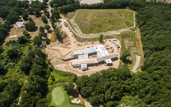 Aerial view of a construction site surrounded by trees and a grassy field. The site includes multiple buildings in various stages of construction, with vehicles and equipment visible. A golf green is in the bottom left corner.