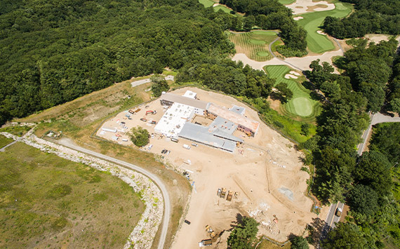 Aerial view of a construction site surrounded by dense trees. The site features multiple buildings under construction. Adjacent to the site is a golf course with several putting greens and fairways. A curved road leads to the area.