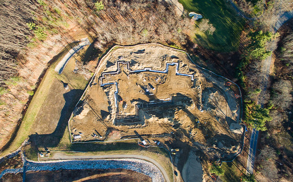 Aerial view of a large construction site with partially built structures surrounded by trees and greenery. The site is marked by dirt, pathways, and equipment, and is bordered by paved and gravel roads.