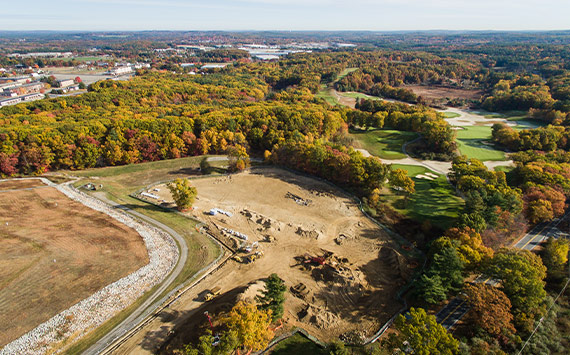 Aerial view of a construction site surrounded by dense forest in autumn colors. Various earth-moving vehicles operate on the sandy area. In the background, a winding road and a golf course with neatly manicured grass are visible.
