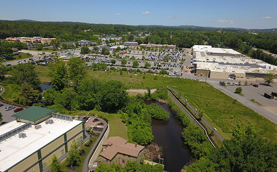 Aerial view of a shopping area surrounded by trees and greenery. Parking lots with numerous cars are visible near several large retail buildings. A small body of water and green fields are in the foreground, extending to forested areas in the distance.