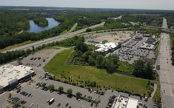 Aerial view of a commercial area near a river. The image shows roads, parking lots, and buildings surrounded by greenery. A river runs parallel to a highway with a mix of forested and open areas in the distance.