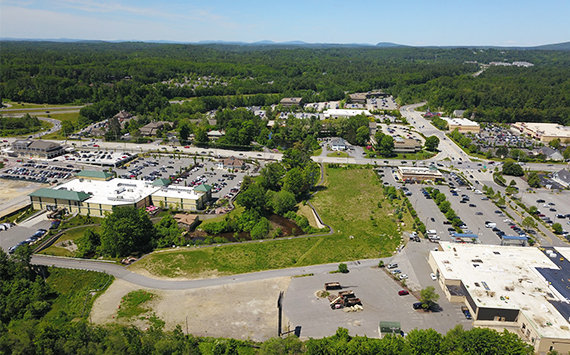 Aerial view of a commercial area surrounded by lush green trees. Several buildings with parking lots are visible. Roads connect the complex, and distant hills are seen on the horizon under a clear blue sky.