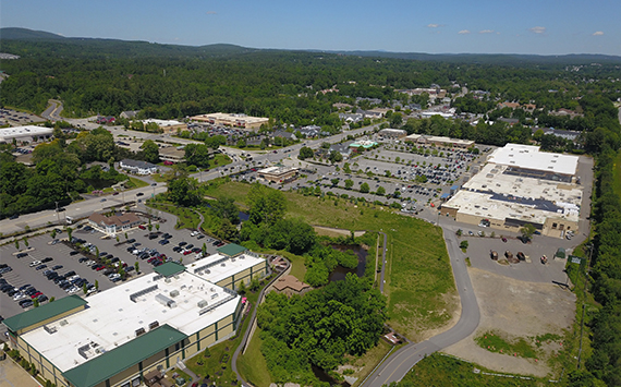 Aerial view of a suburban commercial area with large white-roofed buildings, parking lots, and surrounding greenery. Roads intersect, leading to small wooded areas, with hills visible in the background under a clear blue sky.