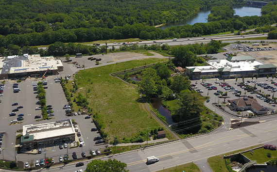 Aerial view of a commercial area featuring parking lots, several buildings, and a grassy central area with a small pond. Trees line the background, and a highway runs parallel to a river in the distance.