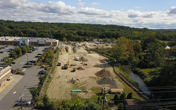 Aerial view of a construction site near a shopping area. Several construction vehicles and equipment are visible on the dirt terrain. A small stream runs parallel to the site, with a backdrop of lush green trees under a cloudy sky.