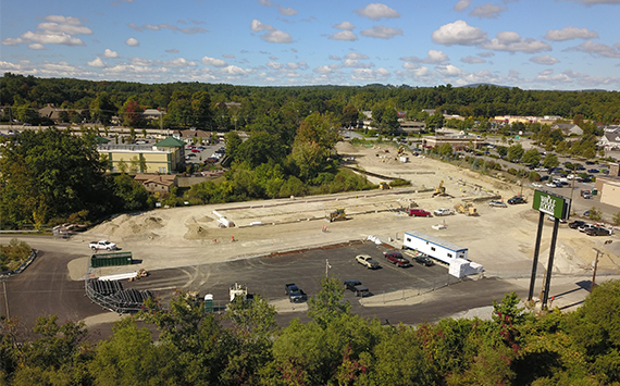 Aerial view of a construction site with heavy machinery, surrounded by trees and buildings. The site is in progress with some foundation work visible. Cars are parked nearby, and a green and white sign is seen on the right. Clouds dot the blue sky.