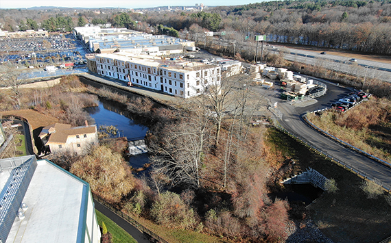Aerial view of an industrial area with several large buildings, a small pond, and surrounding trees. A road runs along the right side, and theres a parking lot with numerous cars in the distance. The landscape is set in a late autumn scene.