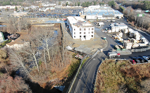 Aerial view of a construction site with a partially built white building at the center. Surrounding the site are stacks of materials, parked vehicles, and nearby trees. In the background, industrial buildings and a parking lot are visible.