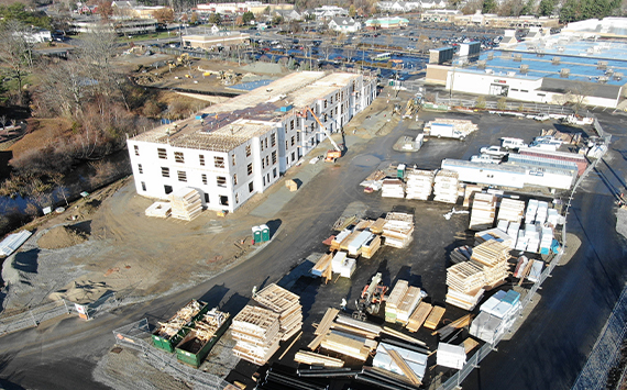 Aerial view of a construction site with a partially-built, multi-story building in the center. Surrounding the structure are stacks of building materials and construction equipment. The area is fenced, and nearby are industrial buildings and trees.