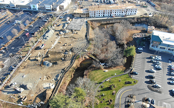 Aerial view of a construction site next to a parking lot and shopping area. The image shows various construction materials and equipment. Trees and a small body of water are visible near the site. Cars are parked in adjacent lots.