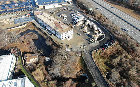 Aerial view of a construction site near a highway. There is a large building under construction surrounded by materials and vehicles. Nearby are trees and a small body of water. Roads and parking areas are visible alongside the site.