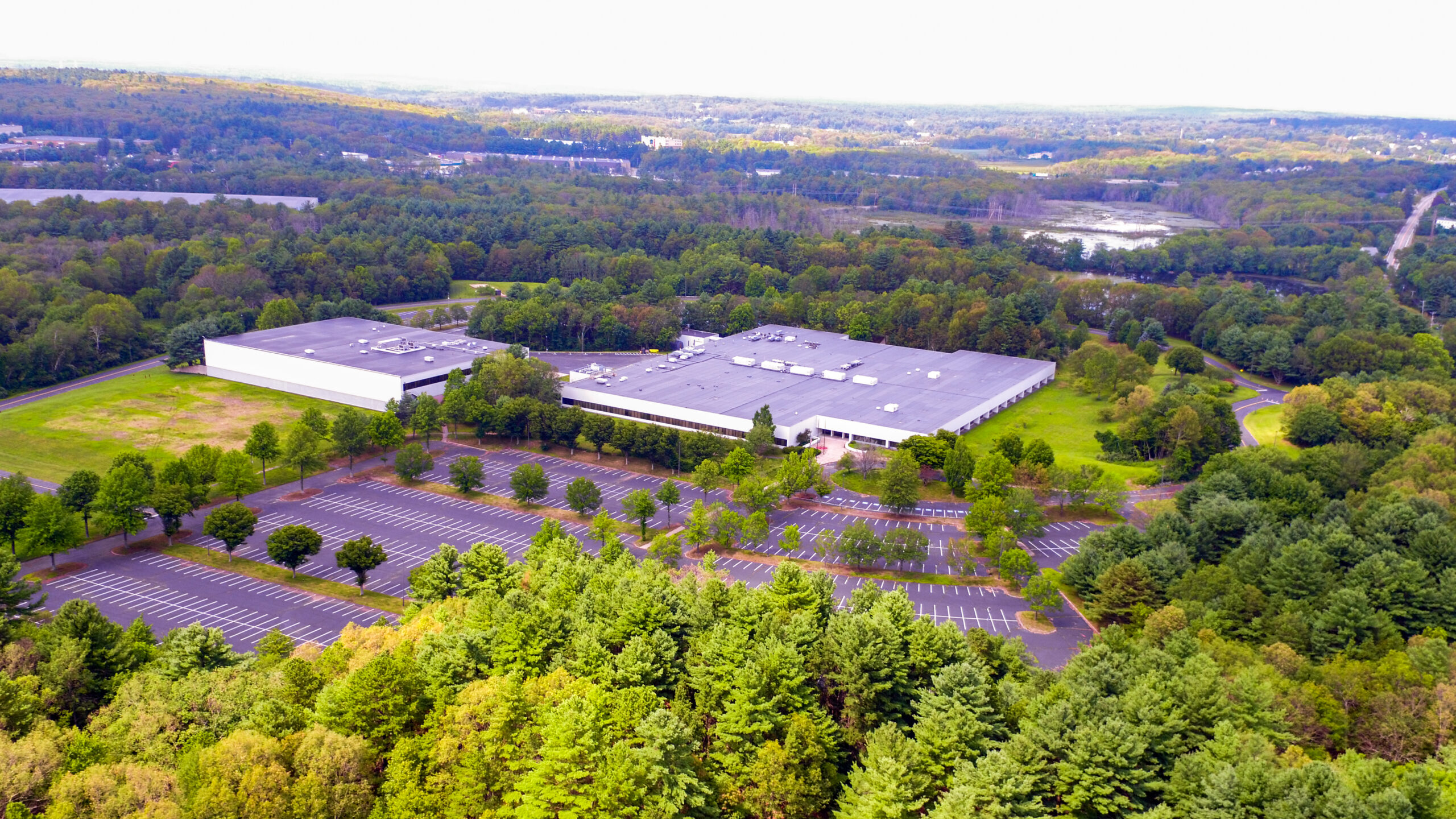 Aerial view of a large industrial building surrounded by dense green forest. The facility has a flat roof and is encircled by a large, mostly empty parking lot. In the background, vast stretches of forest and a road are visible.