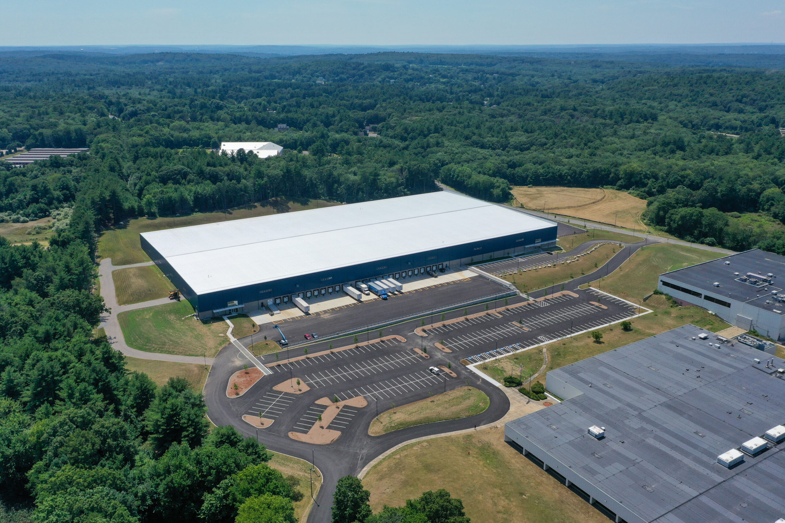 Aerial view of a large warehouse surrounded by parking lots and lush green trees. Several trucks are parked at loading docks. The landscape extends into dense forest with a clear sky above.