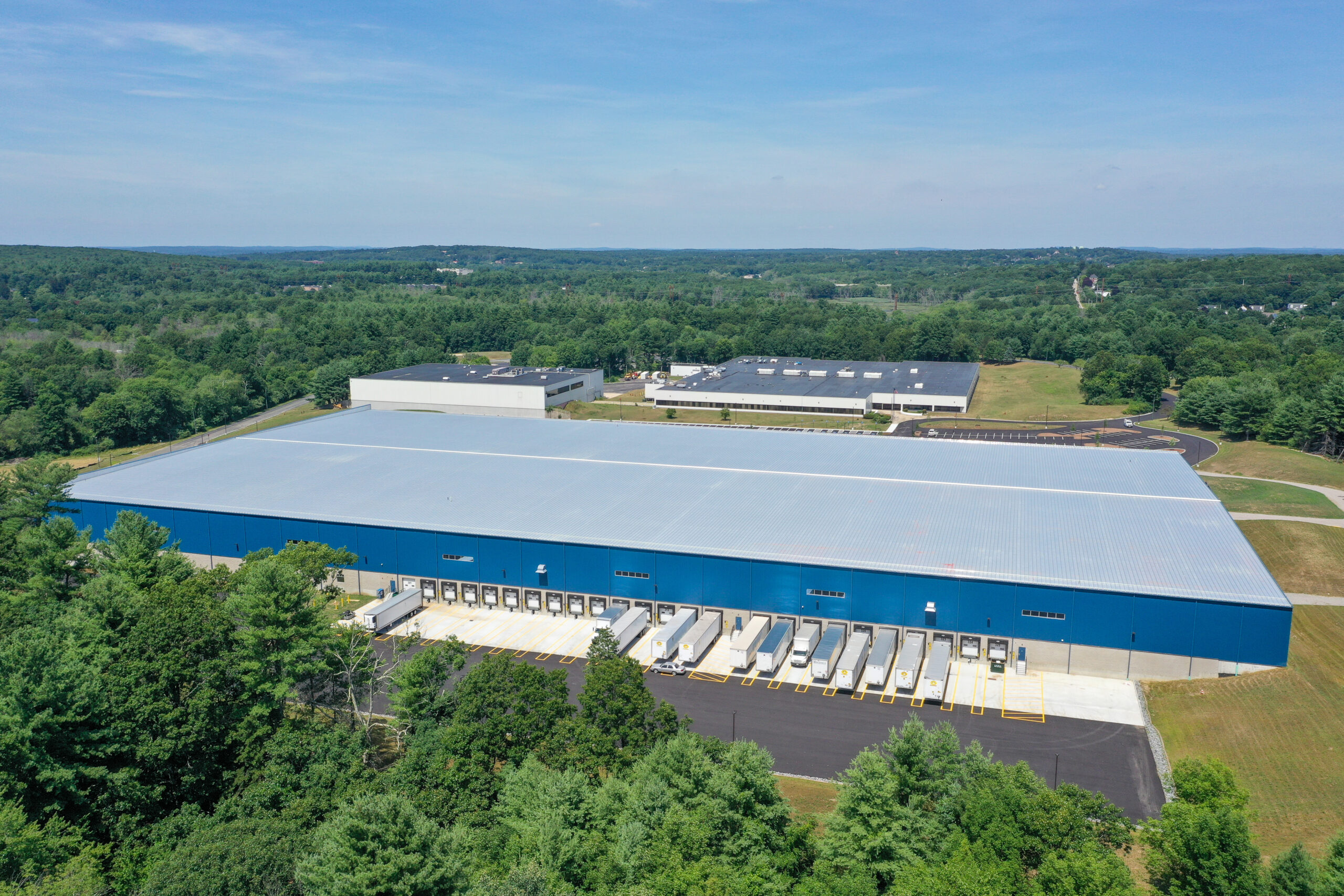 A large blue warehouse with multiple loading docks and several trailers parked outside, surrounded by green trees and vegetation. The warehouse is next to another building, with a sprawling landscape in the background under a clear sky.