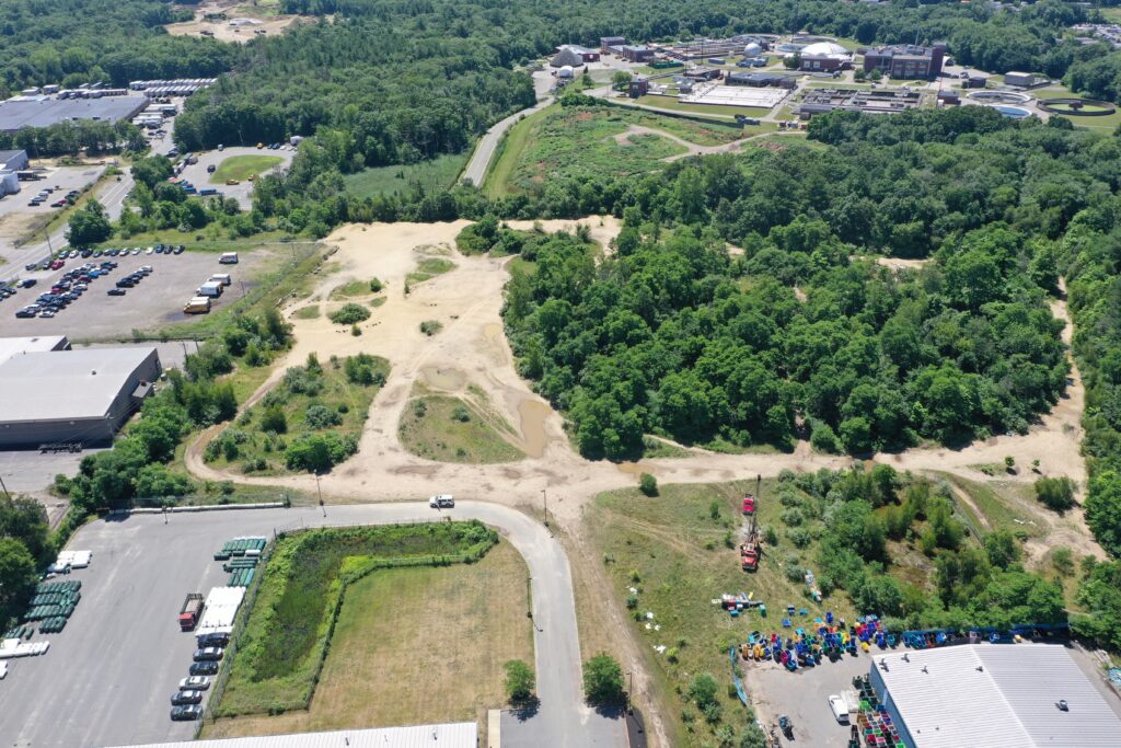 Aerial view of a partially wooded landscape with buildings and parking lots nearby. There are dirt paths and patches of grass, with a collection of colorful objects in the bottom right corner near industrial structures.