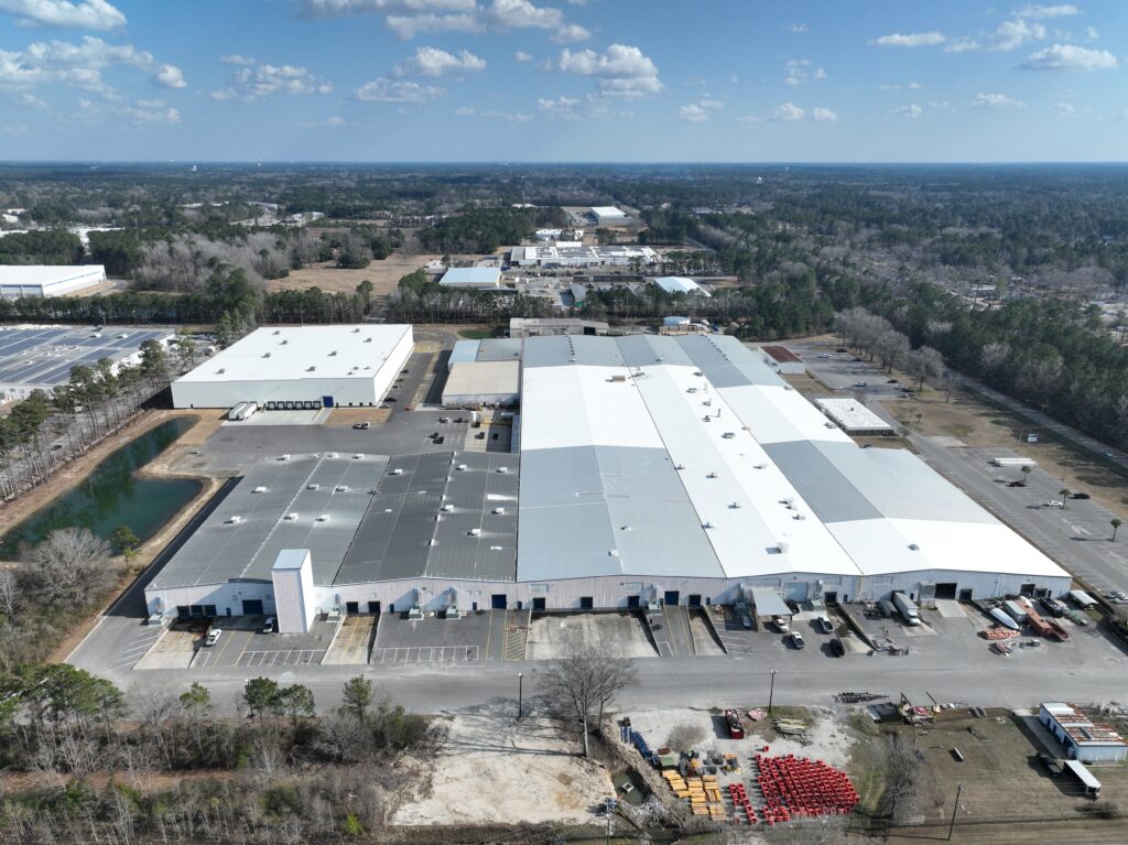 Aerial view of a large industrial complex with multiple warehouses surrounded by trees. The buildings have white and gray roofs, and there are parking areas and open spaces nearby. A small body of water is visible on the left.