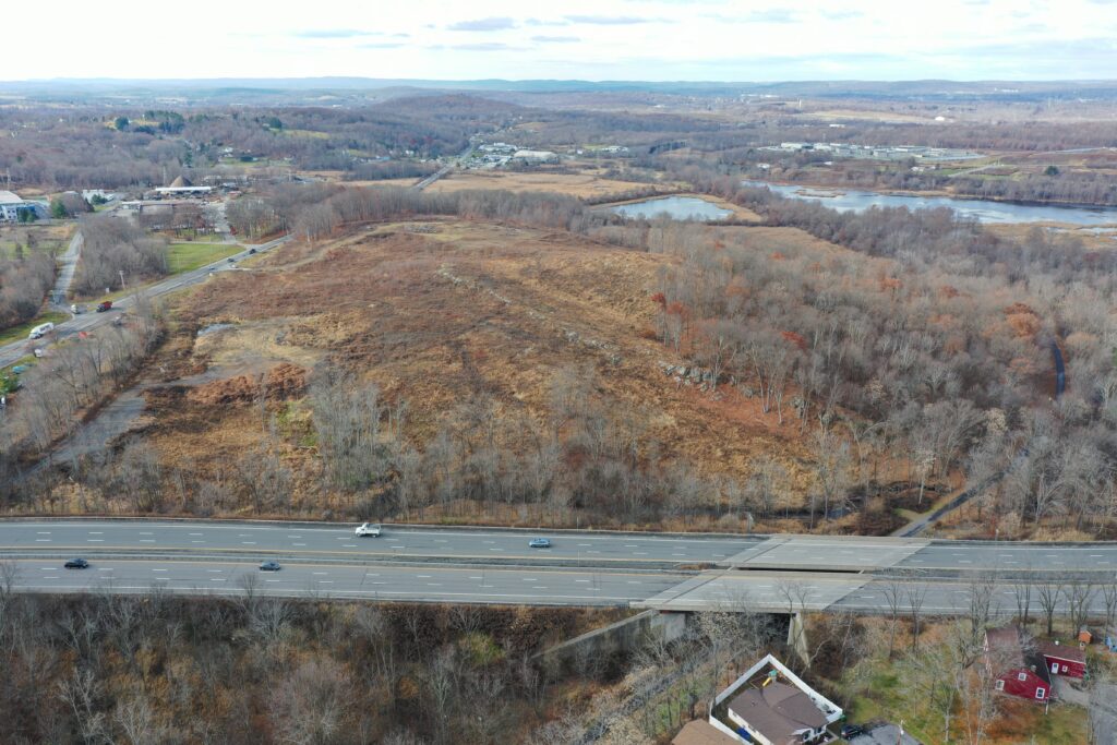 Aerial view of an autumn landscape with a highway cutting through foreground fields and wooded areas. Leafless trees surround a hill and a pond in the background. Sparse buildings are visible, with overcast skies above.