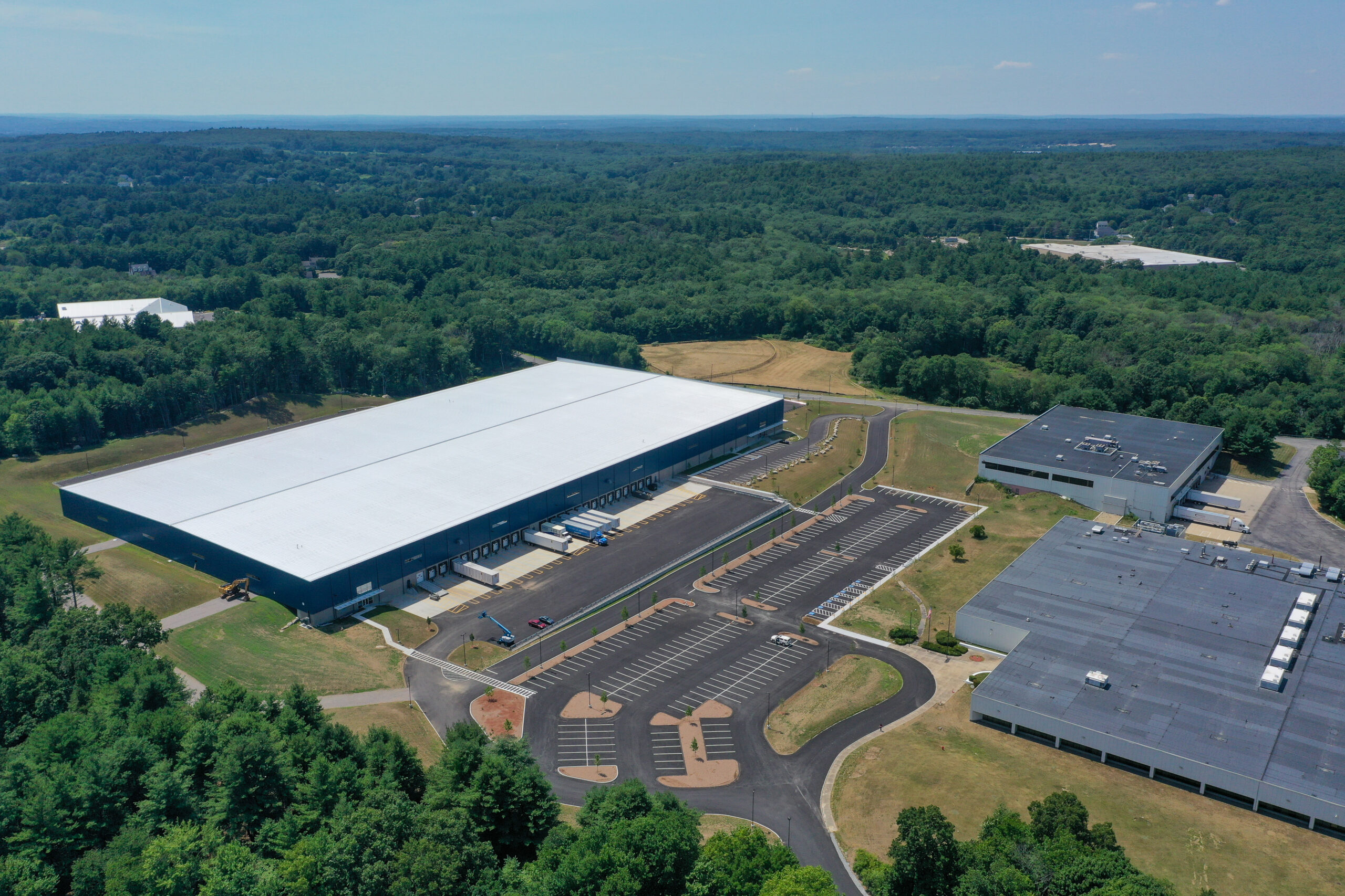 Aerial view of a large industrial complex surrounded by dense greenery. The complex features two main buildings with large parking lots and access roads. The area is nestled in a lush, forested landscape under a clear blue sky.