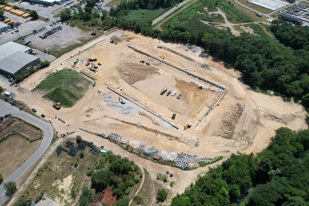 Aerial view of a large construction site surrounded by trees and roads. The site features several earth-moving vehicles, piles of materials, and partially excavated areas. Nearby buildings and parking lots are visible in the top left corner.