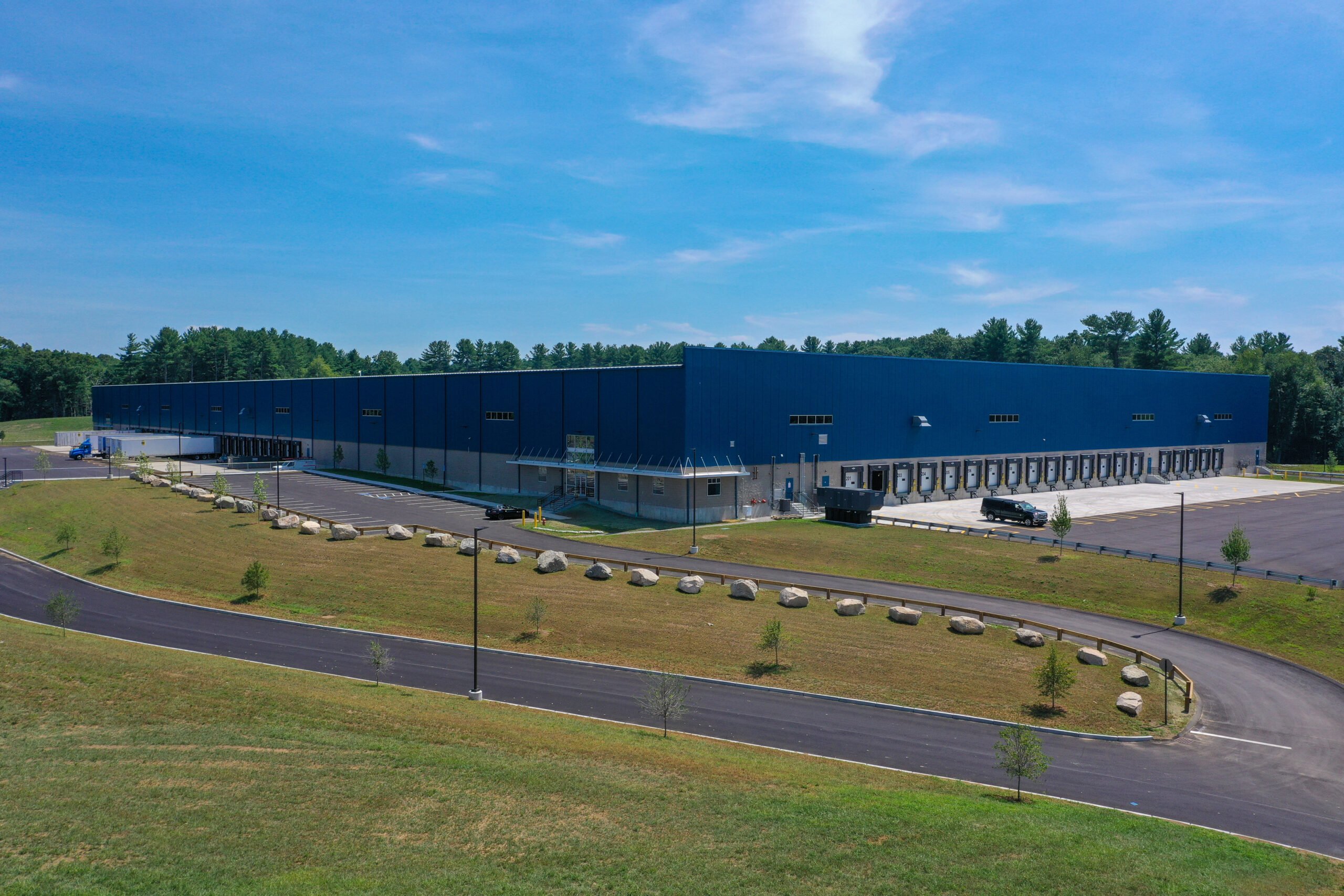 A large blue warehouse sits on a spacious plot with a curved driveway and neat landscaping. The sky is clear and blue, and there are trees in the background. Several loading bays are visible along the side of the building.