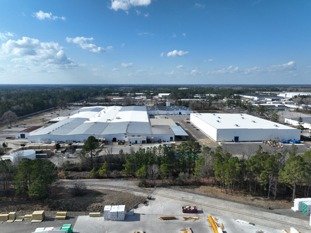 Aerial view of an industrial complex featuring large white warehouse buildings surrounded by trees. The foreground includes a dirt road with scattered material stacks, under a clear blue sky with a few clouds.