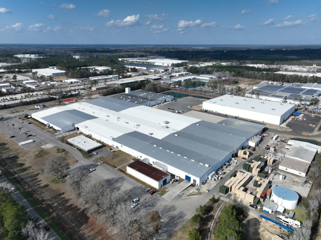 Aerial view of a large industrial complex with white and gray roofs surrounded by trees and roads. Several smaller buildings and parked vehicles are visible around the main structure. The background features more buildings and a forested area.