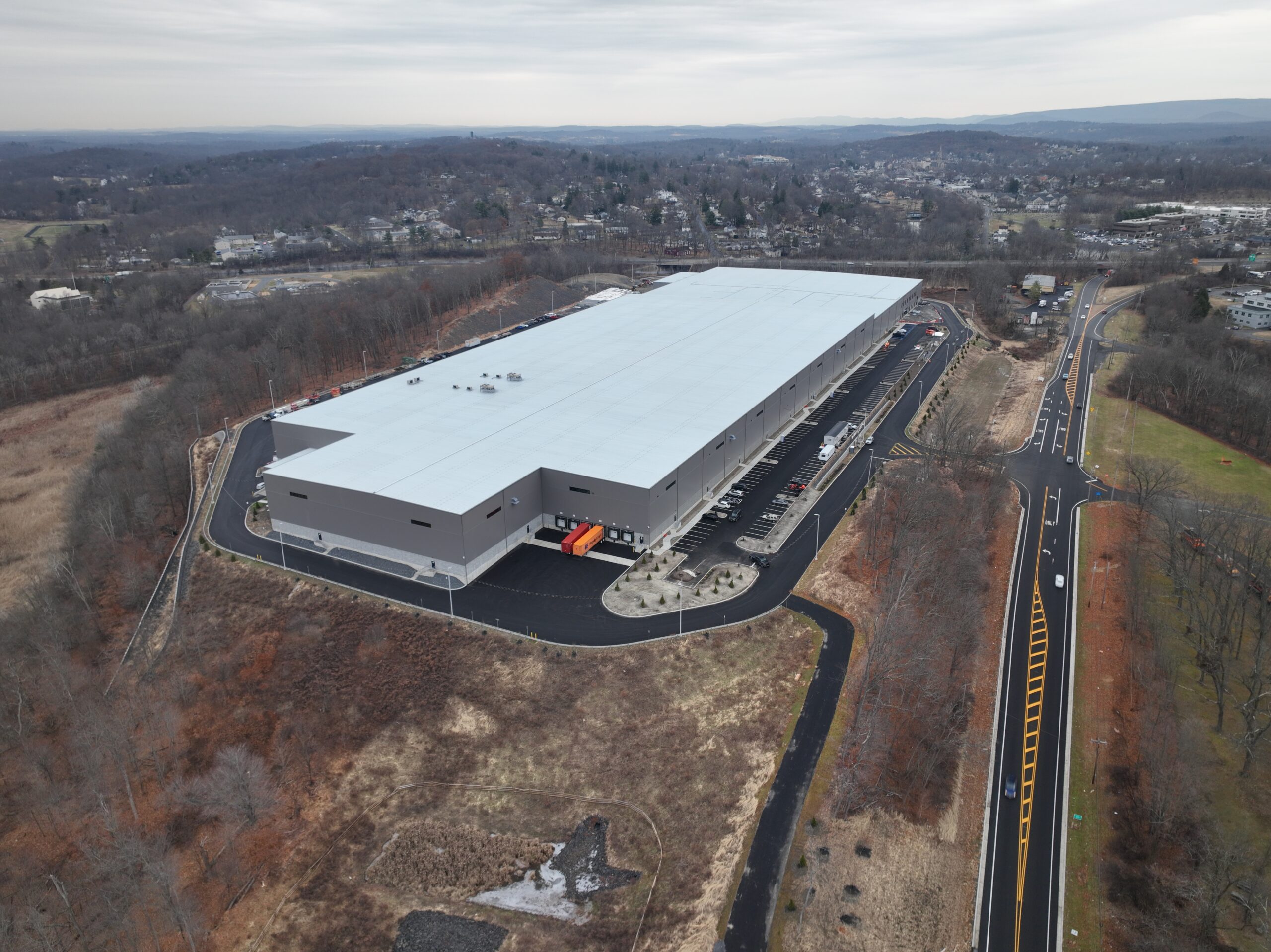 An aerial view of a large, rectangular industrial warehouse with a white roof, situated in a rural area surrounded by trees and roads. The landscape is mostly brown, indicating winter or early spring.