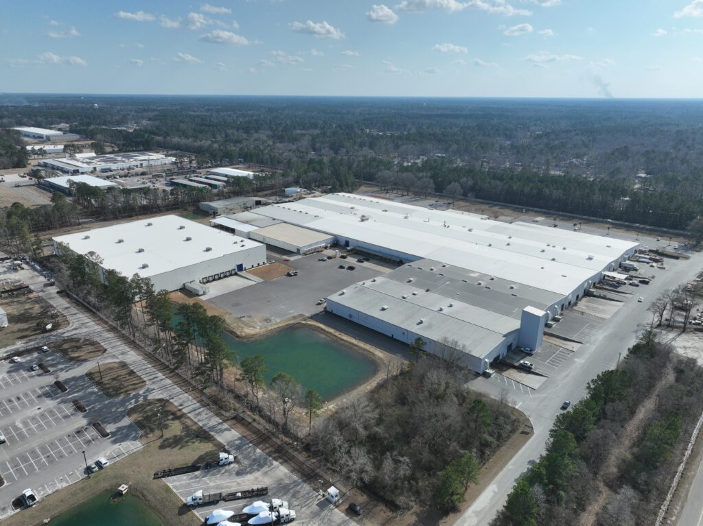 Aerial view of a large industrial facility with multiple white-roofed buildings surrounded by parking lots and greenery. A few ponds and trees are visible around the facility with a distant forest and partly cloudy sky in the background.