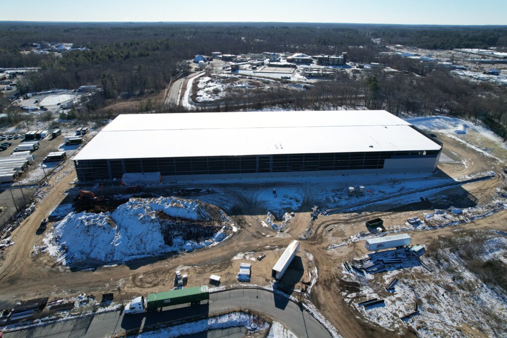 Aerial view of a large, rectangular warehouse under construction, surrounded by dirt and snow-covered ground. Various construction vehicles and materials are visible around the site, with roads and wooded areas in the background.