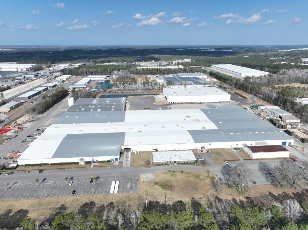 Aerial view of a large industrial facility with multiple connected white-roofed buildings. Surrounding the complex are parking lots, roads, and patches of trees. In the background, more industrial buildings and forested areas are visible under a blue sky.
