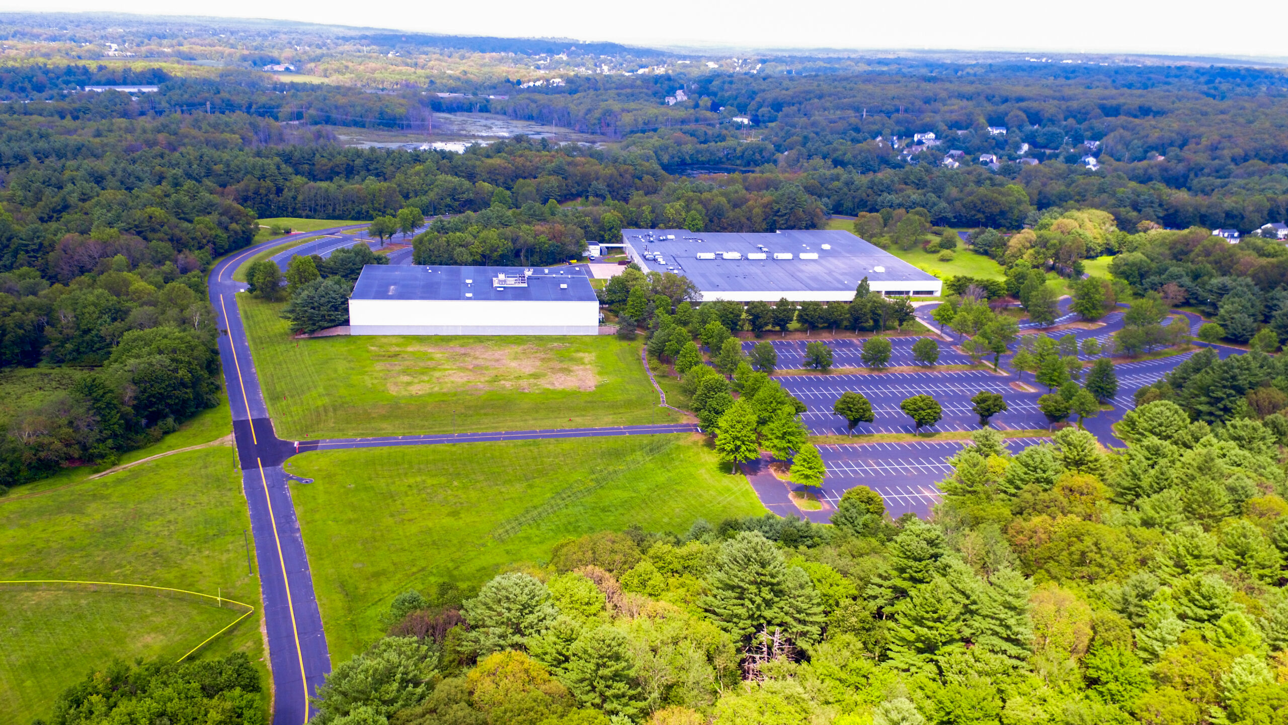Aerial view of a large industrial building surrounded by lush green trees and fields. The parking lot near the building is mostly empty. Roads lead to the facility, and a forested area extends into the distance under a partly cloudy sky.