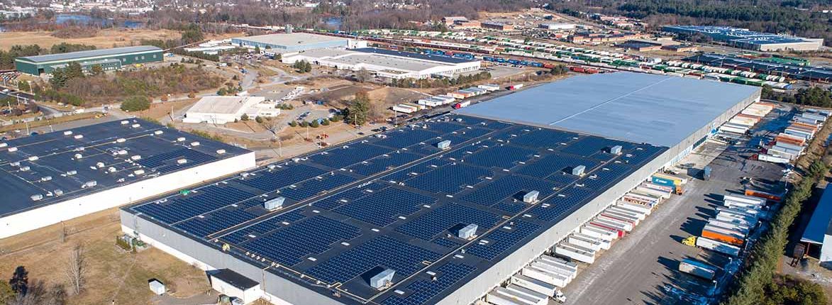 Aerial view of large industrial buildings with solar panels on the roofs, surrounded by parked trucks and trailers. In the background, a railway with several freight cars is visible, along with a wooded area and residential neighborhood.