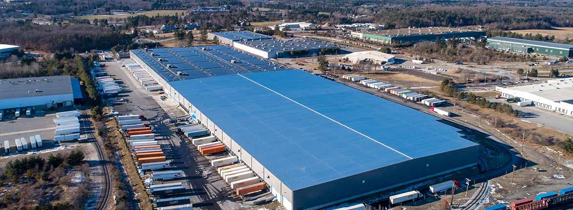 Aerial view of a large industrial warehouse with a blue roof surrounded by numerous parked trailers. The warehouse is situated in a semi-rural area with additional buildings and wooded areas in the background.