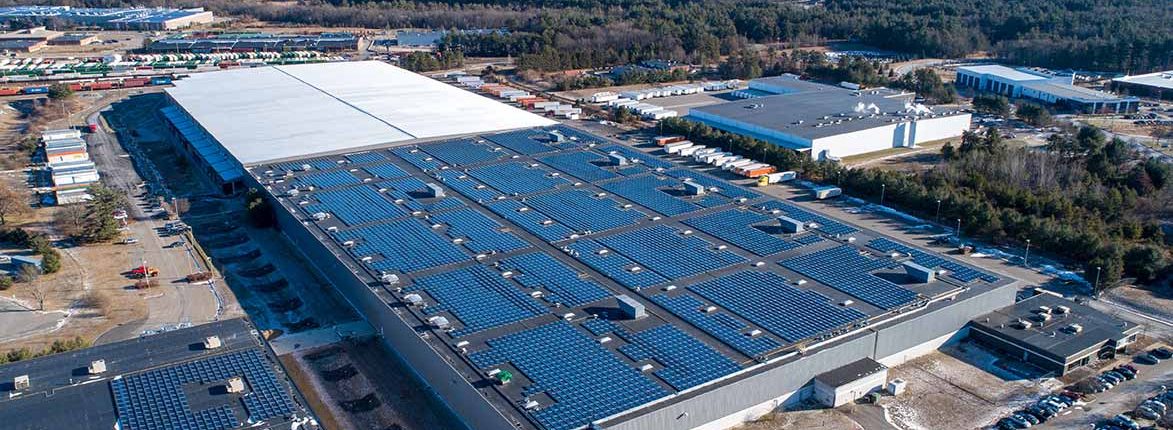 Aerial view of a large warehouse with a roof covered in solar panels. Surrounding the building are various smaller structures and parked trucks. The area is surrounded by trees and industrial buildings, with clear skies in the background.