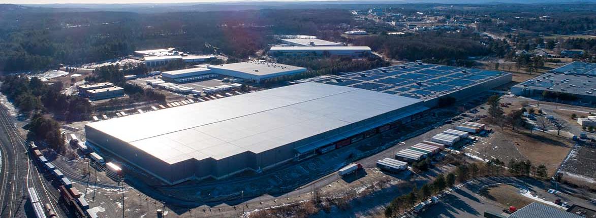 A large industrial warehouse is surrounded by loading docks with several trucks parked outside. The area is expansive, with other warehouses and buildings in the background, bordered by trees under a partly cloudy sky.