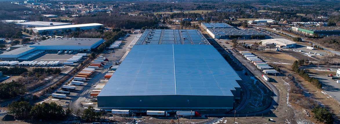 Aerial view of a large rectangular warehouse with numerous semi-trailers parked alongside. Surrounding area includes smaller industrial buildings, trees, and open land. A road is visible in the foreground, and distant hills are in the background.