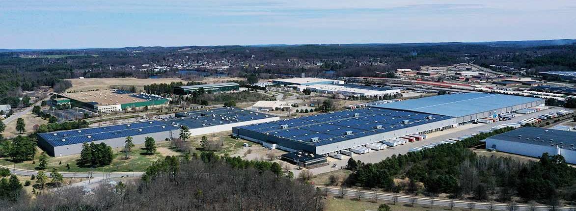 Aerial view of a large industrial complex with multiple warehouses, surrounded by trees and roads. The area features parking lots with trucks and open fields in the background under a clear sky.