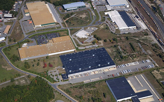 Aerial view of an industrial park with several large warehouse buildings, roads, and parking areas. Surrounding landscape features trees and greenery. A railway line runs along the edge of the park.