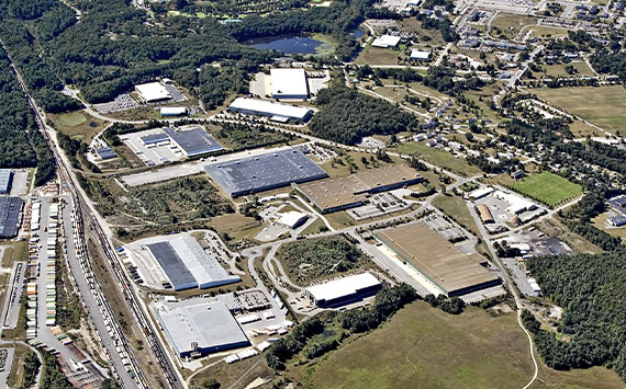Aerial view of an industrial area featuring large warehouses and factories, surrounded by greenery. Roads and rail lines are visible, with residential areas in the distance. The landscape includes patches of trees and open fields.