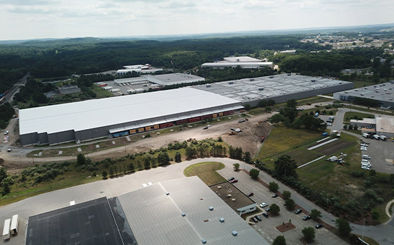 Aerial view of a large industrial complex surrounded by greenery. Multiple expansive warehouse buildings with gray roofs are visible. Roadways, parking lots, and a few parked vehicles are seen around the structures. The sky is cloudy.
