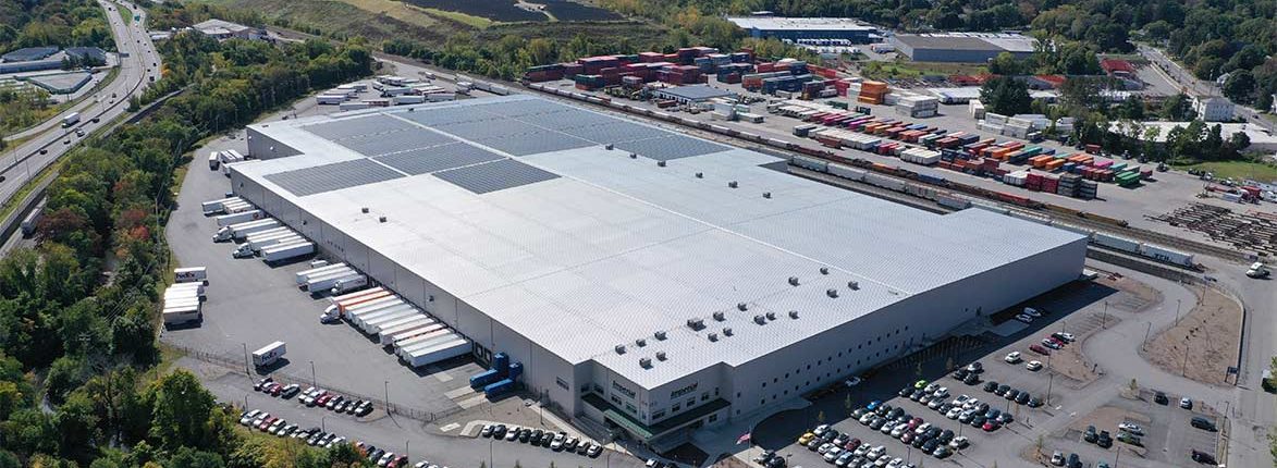 Aerial view of a large warehouse with a white roof and solar panels. Surrounding the warehouse are numerous parked trucks and shipping containers. Nearby greenery and roads are visible, along with other industrial buildings in the background.