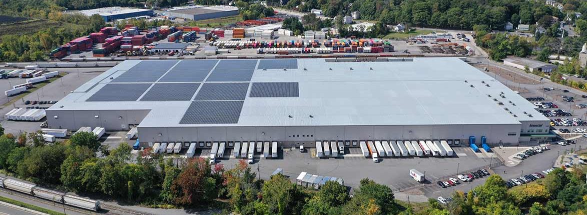 Aerial view of a large warehouse with numerous loading docks and solar panels on the roof. Several trucks are parked at the docks. Surrounding the warehouse are trees, parking areas, and a yard with stacks of shipping containers.