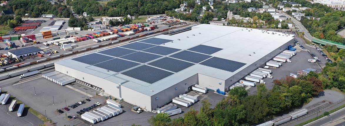 Aerial view of a large industrial warehouse with solar panels on the roof. The facility is surrounded by trucks, trailers, and a railway line. Green trees and a few smaller buildings are visible in the background.