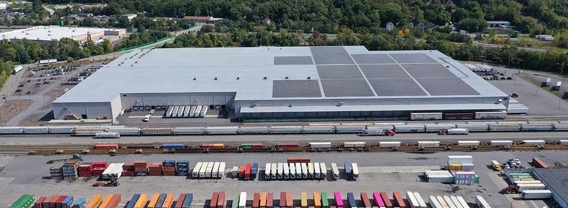 Aerial view of a large industrial warehouse with a gray roof, surrounded by greenery. Numerous trucks, trailers, and shipping containers are lined up in a yard next to railway tracks, indicating transportation and logistics activity.